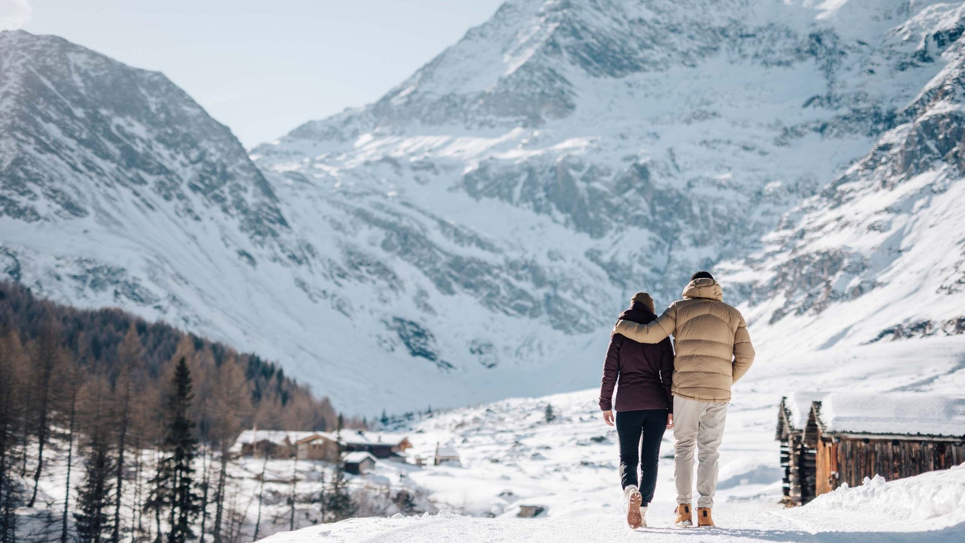 Paar spaziert Arm in Arm im verschneiten Bergdorf mit schneebedeckten Bergen